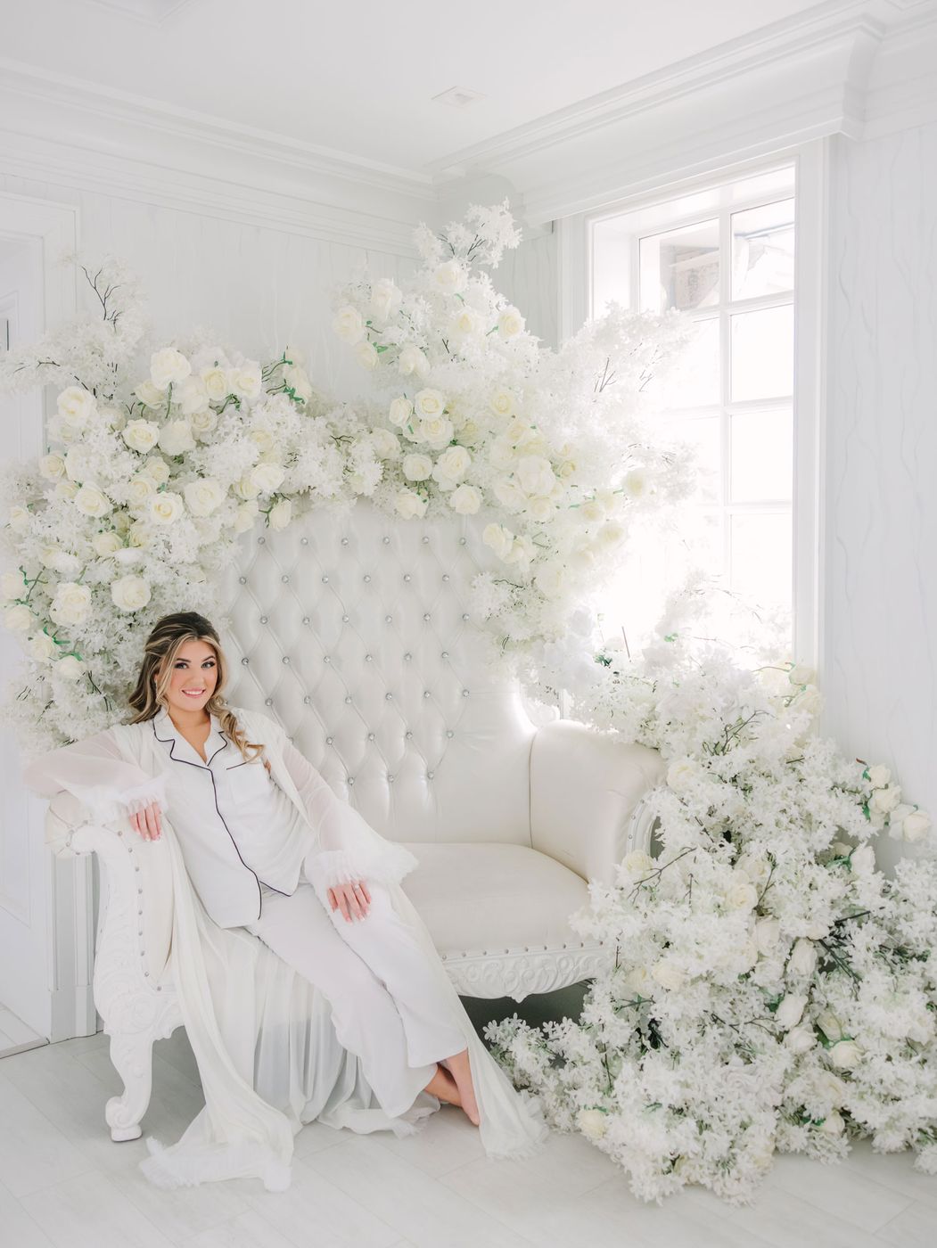 Bride in a white robe seated on a tufted chair framed by white flowers