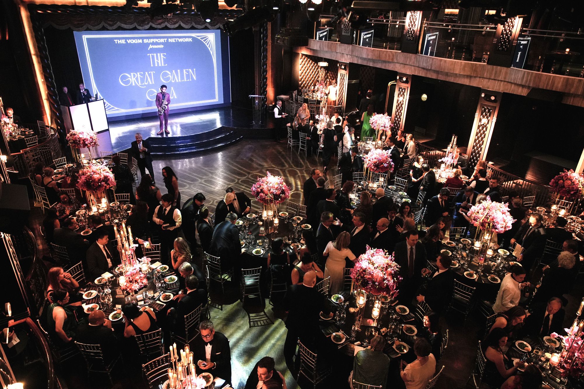 Guests at candlelit tables with tall pink centerpieces, seen from the balcony, with the gala name on the stage screen