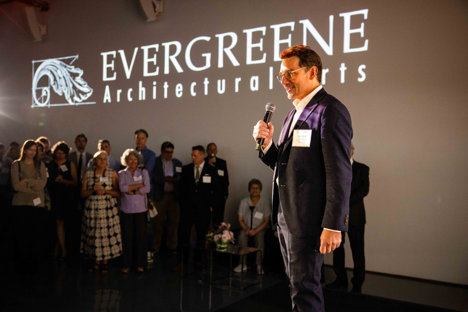 Speaker with a microphone in front of the Evergreene Architectural Arts logo, guests standing behind him