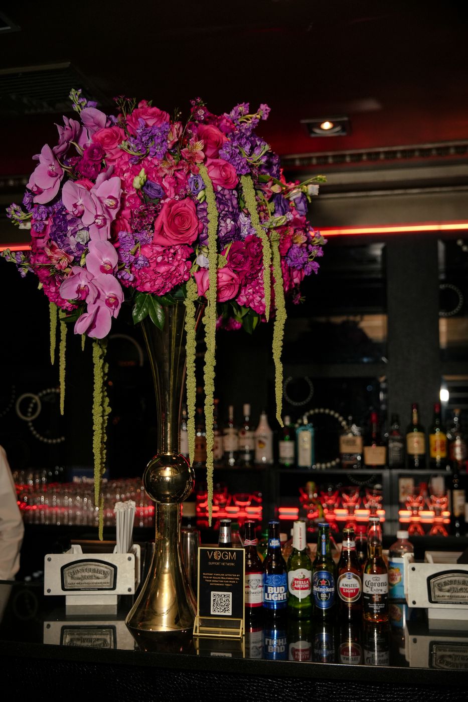 Tall arrangement of pink orchids, roses and hanging amaranthus in a gold vase on the bar