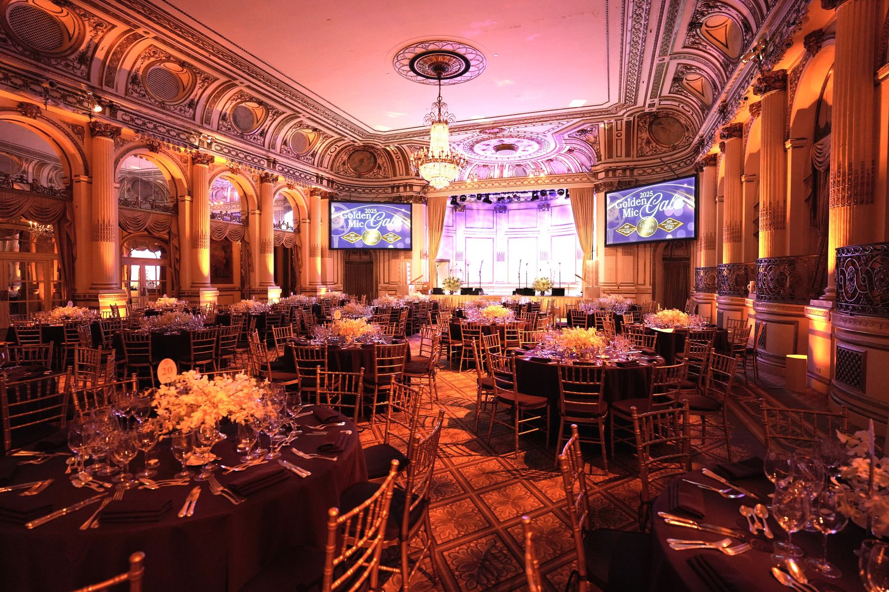 The Plaza Grand Ballroom set for a gala, with gold chairs, chandeliers and a stage between two screens