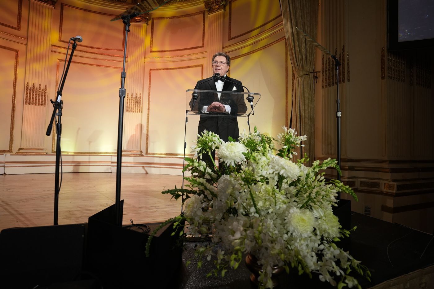 Speaker in a tuxedo at a clear podium behind a large white flower arrangement