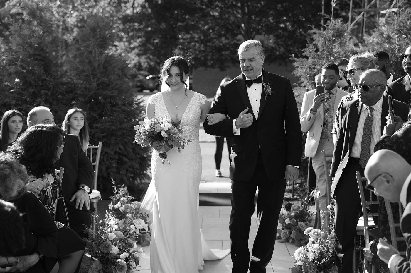 Black and white photo of the bride being escorted down an outdoor aisle