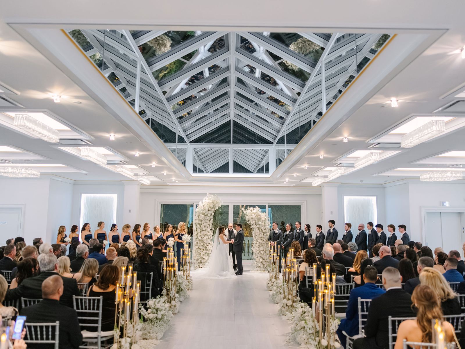 Wedding ceremony under a glass roof with a candlelit aisle and a floral arch
