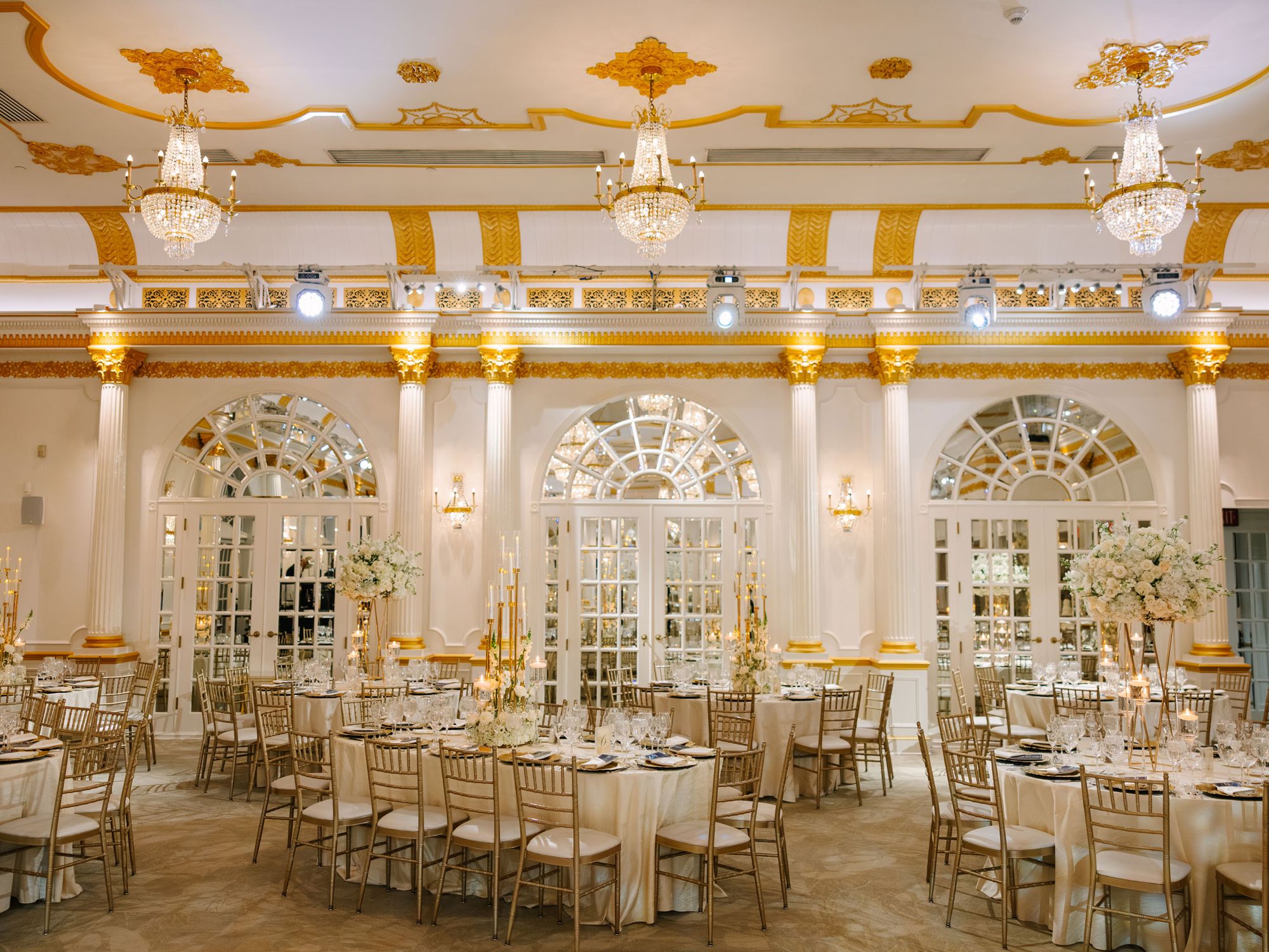 Gold and white ballroom with crystal chandeliers set for a wedding reception