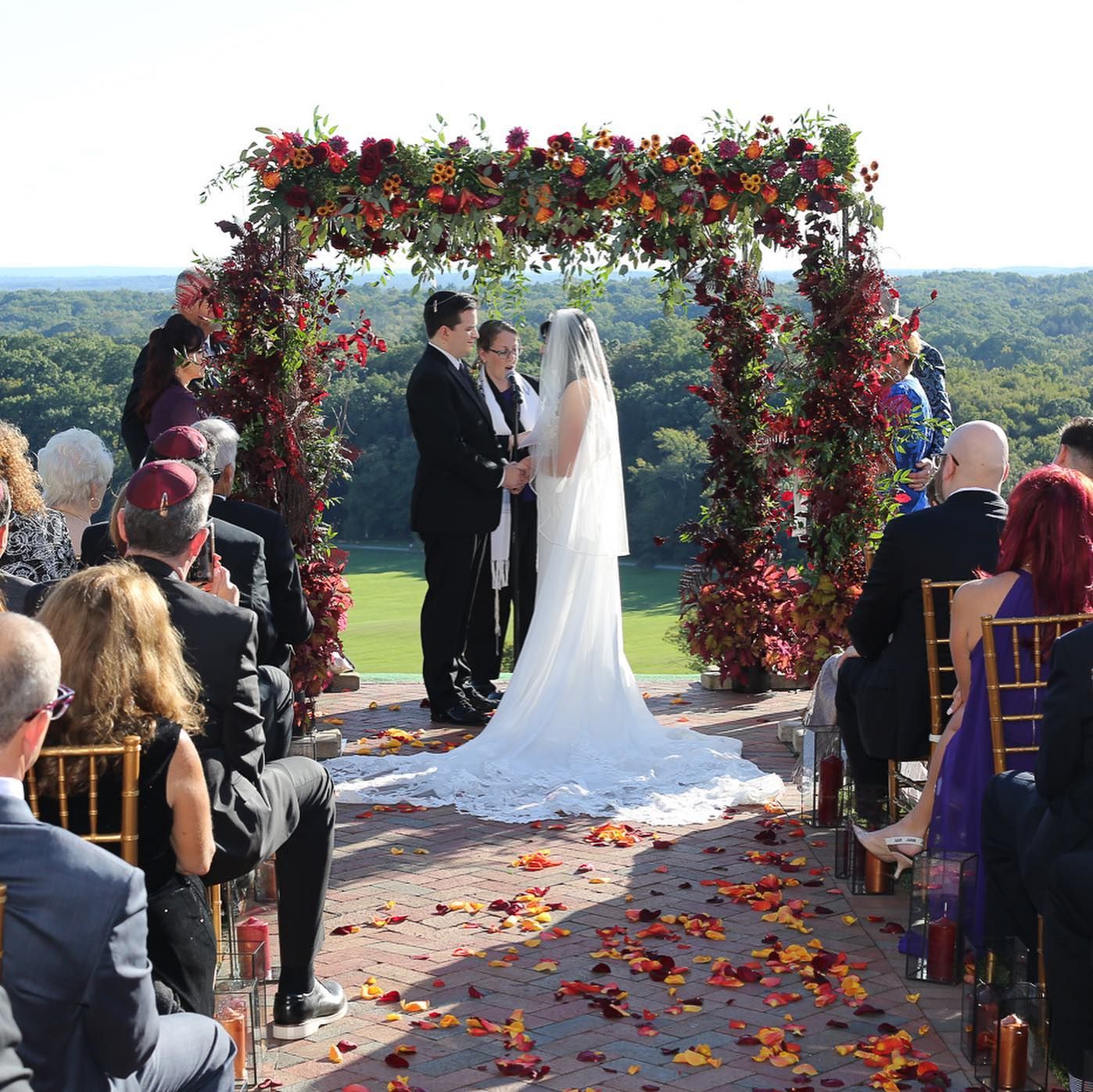 Wedding ceremony under an arch of autumn flowers with wooded hills behind